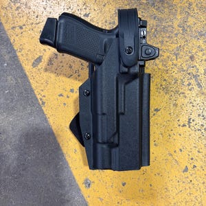 May include: Black handgun in a black holster. The handgun is positioned at an angle, with the barrel and grip visible. The holster has a molded design and is secured with screws. The background is a yellow and gray surface.