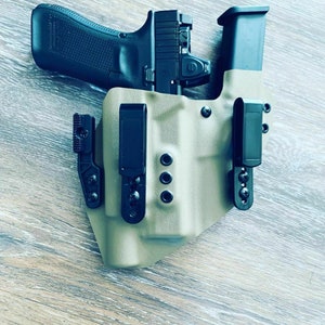 May include: A tan leather holster with two black clips and a black handgun inside. The holster has a textured surface and is designed to be worn on a belt.