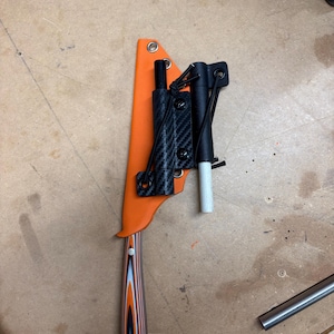 May include: An orange knife sheath with a black and carbon fiber attachment. The knife handle has a layered wood design in shades of orange, black, and white. A black cylindrical object and a white cylindrical object are attached to the sheath.