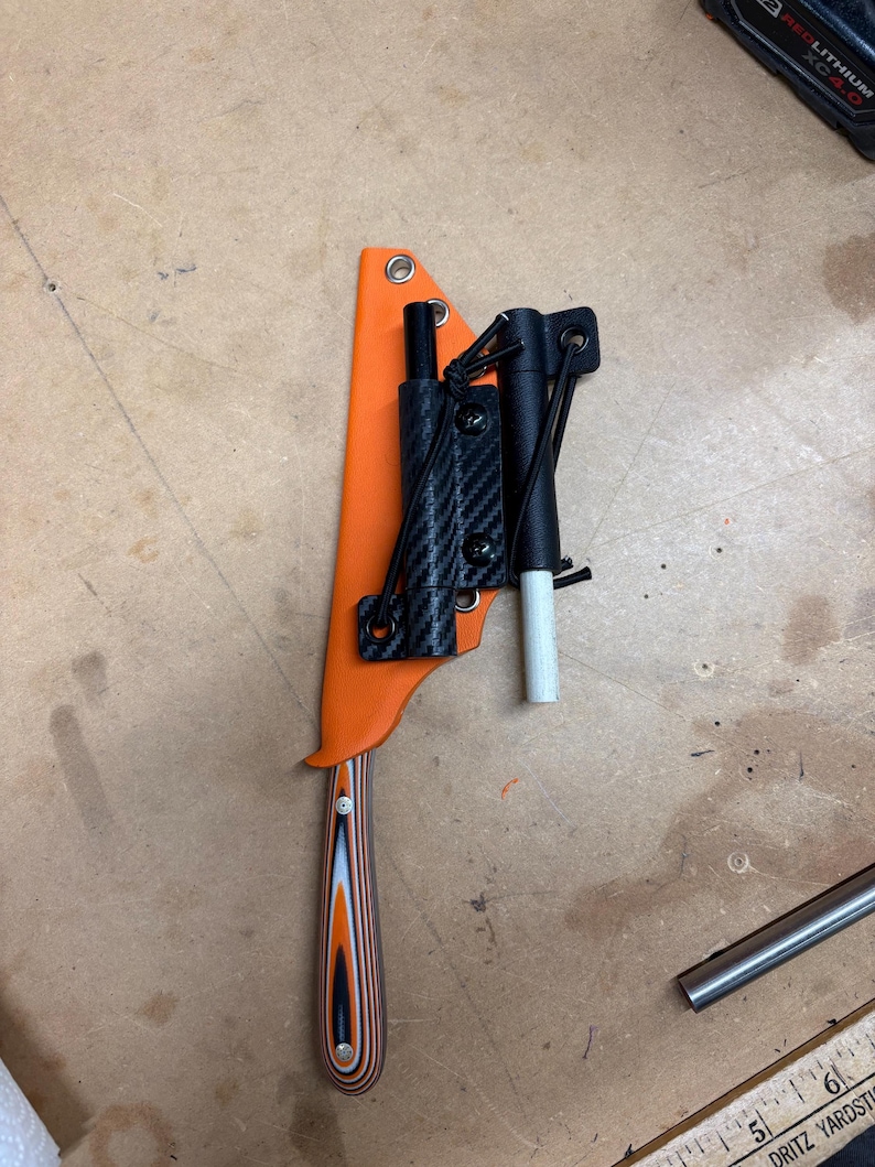 May include: An orange knife sheath with a black and carbon fiber attachment. The knife handle has a layered wood design in shades of orange, black, and white. A black cylindrical object and a white cylindrical object are attached to the sheath.