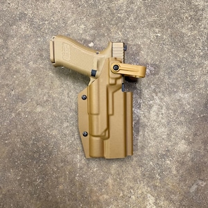 May include: Tan-colored handgun in a matching tan holster. The handgun is positioned inside the holster, with the barrel facing left. The holster has a belt clip. The background is a textured, neutral-toned surface.