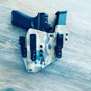 May include: A camouflage-patterned holster with two black clips and a black magazine holder. A black handgun with a red laser sight is partially visible inside the holster.