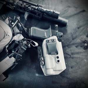 Puede incluir: Un surtido de equipo táctico, que incluye un arma de fuego con una óptica montada, un rifle equipado con luz y una funda. Los artículos están dispuestos sobre una superficie oscura y texturizada, con una combinación de colores monocromática.