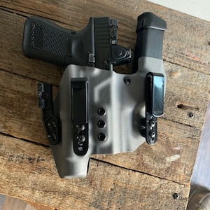 May include: A black handgun with a red dot sight is holstered in a grey and black tactical holster. The holster has two black clips and is resting on a wooden surface.