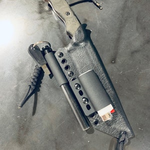 Puede incluir: Una funda de cuero negra con un cuchillo, un iniciador de fuego y un encendedor en su interior. La funda tiene una correa de paracord negra y un anillo de metal.