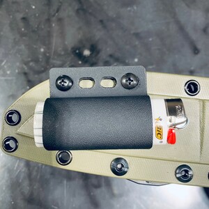 Puede incluir: Un porta mechero negro y gris con un mechero negro unido a un cuchillo verde. El porta mechero tiene dos tornillos a cada lado y tres agujeros en el medio. El mechero es un mechero BIC con un dise&ntilde;o rojo y amarillo.