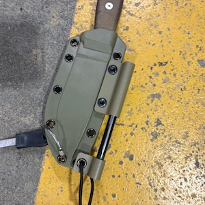 May include: An olive green knife sheath with a black handle and a brown strap. The sheath has a black cord and a metal piece attached. The sheath is secured with black screws. The background is a yellow and gray surface.