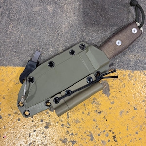 May include: An olive green knife sheath with a brown handle and a black cord. The sheath has a small, cylindrical attachment and a metal piece on the side. The knife is designed for outdoor use.