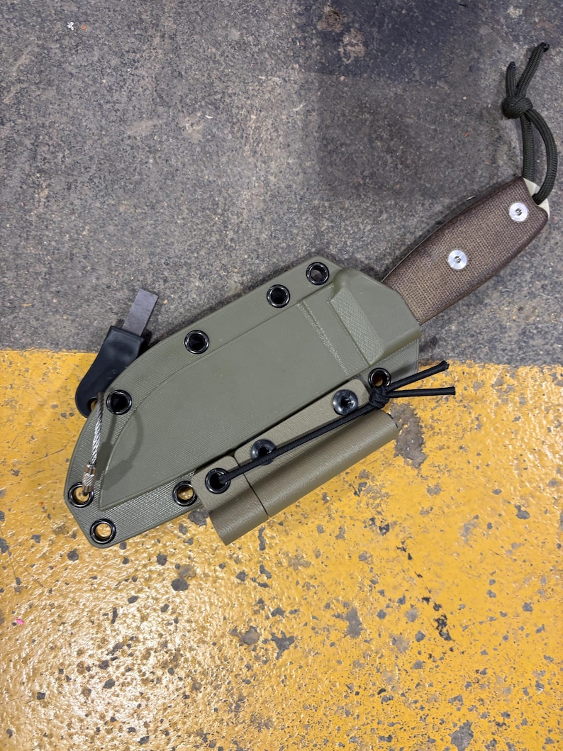 May include: An olive green knife sheath with a brown handle and a black cord. The sheath has a small, cylindrical attachment and a metal piece on the side. The knife is designed for outdoor use.