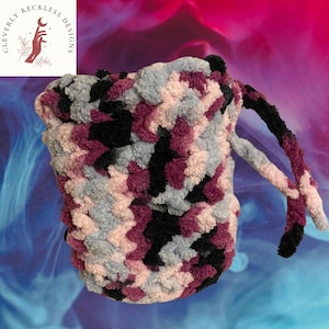 May include: A handmade, plush crochet bag in a mix of colors including black, gray, and shades of purple. The bag has a drawstring closure and a matching handle. The bag is made with a soft, textured yarn, and the design is a unique, eye-catching pattern.