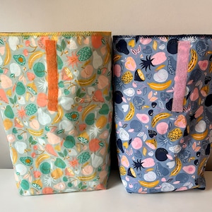 Peut inclure: Deux bacs de rangement en tissu avec un motif imprimé de fruits. Le bac de gauche est vert avec un imprimé de pêche, d'avocat et de banane. Le bac de droite est bleu avec un imprimé de fruits rose, jaune et bleu.