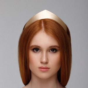 May include: A woman with long, straight, red hair wearing a gold crown headband. She is wearing a light brown v-neck shirt.