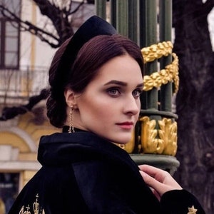 May include: A woman with dark brown hair wearing a black velvet headband, a black velvet jacket with gold embroidery, and gold earrings. She is standing in front of a gold and green decorative pillar.