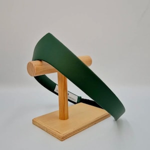 May include: A dark green headband displayed on a wooden stand. The headband has a wide, flat design and a smooth texture. The stand is made of light-colored wood, with a rectangular base and a curved support.