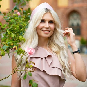 May include: A woman wearing a light pink, one-shoulder dress with a ruffled neckline and a matching hat. She is smiling, with long blonde hair and a black wristwatch. Pink roses and green foliage are in the foreground.