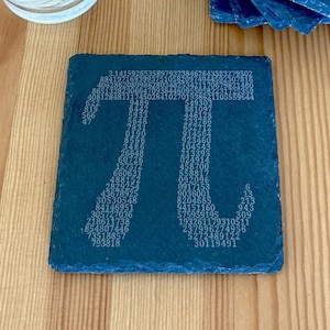 Puede incluir: Un posavasos de pizarra negra con la constante matemática pi representada por los dígitos de pi. El posavasos está sobre una superficie de madera con un vaso de agua y una pequeña planta en maceta.