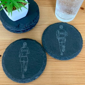 May include: Dark grey slate coasters featuring etched running figures, with space for a name on the jersey. A small green plant in a white pot and a glass of water are also visible in the image.