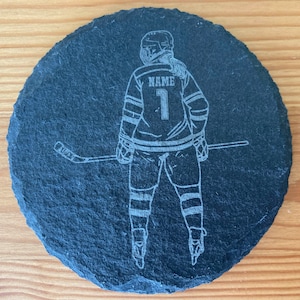 Puede incluir: Posavasos de pizarra negra con una silueta blanca de un jugador de hockey sosteniendo un palo. El jugador lleva una camiseta con la palabra "NAME" y el n&uacute;mero "1" en la espalda.