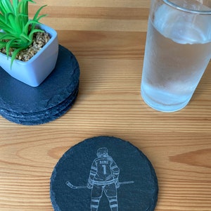 Puede incluir: Posavasos de pizarra negra con un dise&ntilde;o grabado en blanco de un jugador de hockey sosteniendo un palo. La camiseta del jugador tiene la palabra "NAME" y el n&uacute;mero "1" en la espalda.