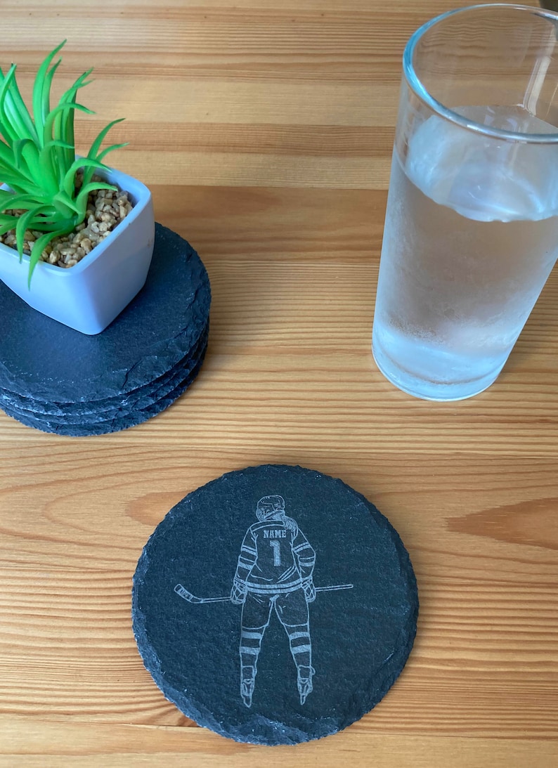 Puede incluir: Posavasos de pizarra negra con un dise&ntilde;o grabado en blanco de un jugador de hockey sosteniendo un palo. La camiseta del jugador tiene la palabra "NAME" y el n&uacute;mero "1" en la espalda.