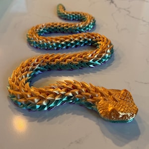 Può includere: Una figura di serpente articolata stampata in 3D con un corpo segmentato e una finitura metallica. Le squame presentano una sfumatura di colore che va da un oro vibrante sulla parte superiore a un verde smeraldo intenso e viola sui lati.
