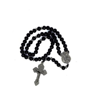 May include: A black rosary with round beads and a silver-colored crucifix. The crucifix features a detailed depiction of Jesus Christ. The rosary also includes a silver-colored shield-shaped pendant.