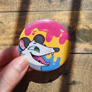 May include: A circular pin-back button featuring a cartoon opossum with white fur, a black nose, and green eyes. The background displays a vibrant pattern of pink, yellow, and blue paint drips with a subtle holographic finish across the surface.