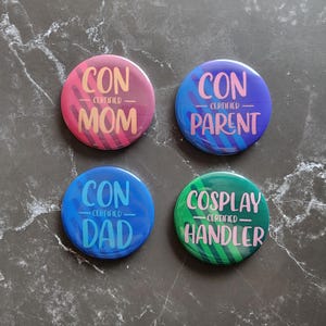 May include: Four circular pin-back buttons with diagonal striped backgrounds. The pink button reads CON CERTIFIED MOM, the purple button reads CON CERTIFIED PARENT, the blue button reads CON CERTIFIED DAD, and the green button reads COSPLAY CERTIFIED HANDLER.