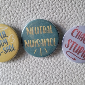 May include: Three round buttons with different colored backgrounds and gold lettering. The first button is yellow with a halo and wings and says "Lawful Goody Two-Shoe". The second button is teal with gold lettering that says "Neutral Nuisance". The third button is purple with a red devil tail and says "Chaotic Stupid".
