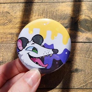 Pode incluir: Um bottom redondo com uma ilustração de desenho animado de um gambá branco com um sorriso largo, olhos verdes e orelhas rosas. O fundo consiste em listras horizontais em amarelo, branco e roxo com um efeito de tinta escorrendo.