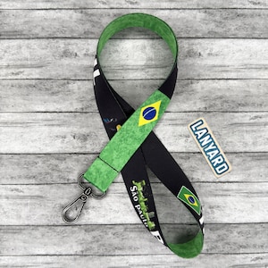May include: Black lanyard with a green and yellow Brazilian flag design. The lanyard has a metal clasp and the words "Sao Paulo" are printed on the black fabric. A small tag that says "LANYARD" is also present.