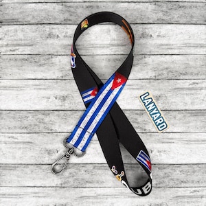 May include: Black lanyard with a silver clasp, featuring a design incorporating the Cuban flag and other graphics. The word "LANYARD" is printed on a small rectangular tag. The lanyard is displayed against a light gray wooden background.
