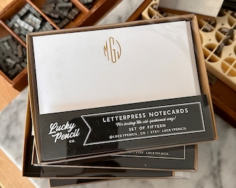 May include: A stack of letterpress notecards in a cardboard box. The cards are white with a gold monogram. The box has a black and white label that reads "LETTERPRESS NOTECARDS" and "Lucky Pencil Co."