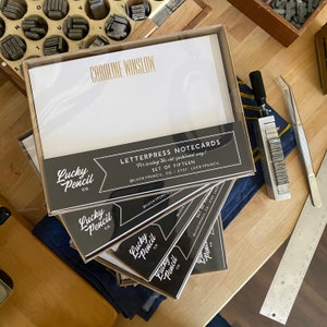 May include: A stack of letterpress notecards in a clear box, with the name "Caroline Winslow" printed in gold. The cards are labeled "Letterpress Notecards" and "Set of Fifteen" with the "Lucky Pencil Co." logo. Other printing tools are in the background.