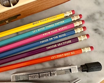 May include: A collection of colorful pencils with motivational phrases printed in gold. The pencils are yellow, teal, blue, pink, purple, and orange. The phrases include "MAKE TODAY COUNT" and "LET THAT SHIT GO." Also included are a metal type holder, tweezers, and a wooden box.