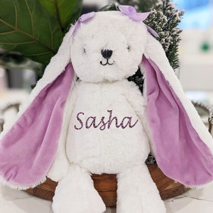 Puede incluir: Un peluche blanco con forma de conejo con largas orejas forradas en lavanda y un lazo a juego. El nombre "Sasha" est&aacute; bordado en el pecho. El conejo est&aacute; colocado sobre una bandeja de madera.