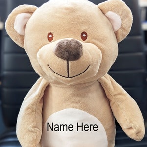 May include: A beige plush teddy bear with a white circle on its chest that says "Name Here". The bear has brown eyes and a brown nose.