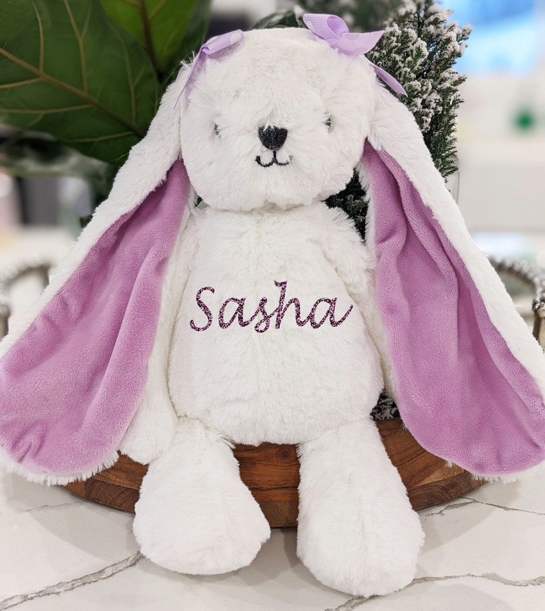 Puede incluir: Un peluche blanco con forma de conejo con largas orejas forradas en lavanda y un lazo a juego. El nombre "Sasha" est&aacute; bordado en el pecho. El conejo est&aacute; colocado sobre una bandeja de madera.