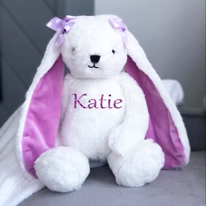 Puede incluir: Un conejo de peluche blanco con orejas largas y ca&iacute;das y un interior morado. El conejo tiene un lazo morado con lunares blancos y el nombre "Katie" bordado en el pecho. Est&aacute; sobre una superficie gris.