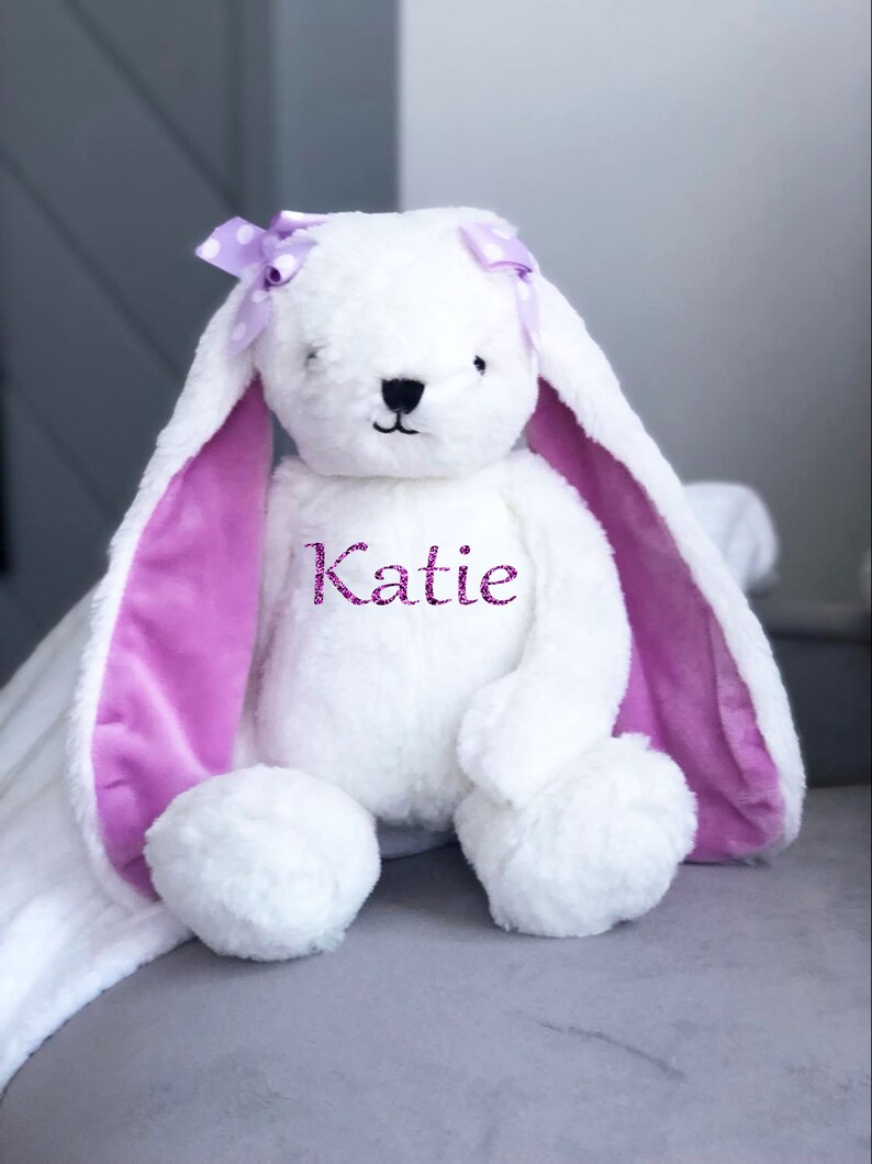 Puede incluir: Un conejo de peluche blanco con orejas largas y ca&iacute;das y un interior morado. El conejo tiene un lazo morado con lunares blancos y el nombre "Katie" bordado en el pecho. Est&aacute; sobre una superficie gris.