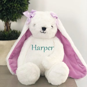 Puede incluir: Un conejito de peluche blanco con orejas largas y un interior morado. El conejito tiene un lazo morado con lunares y el nombre "Harper" bordado en el pecho en color turquesa. El conejito est&aacute; sentado sobre una superficie gris.