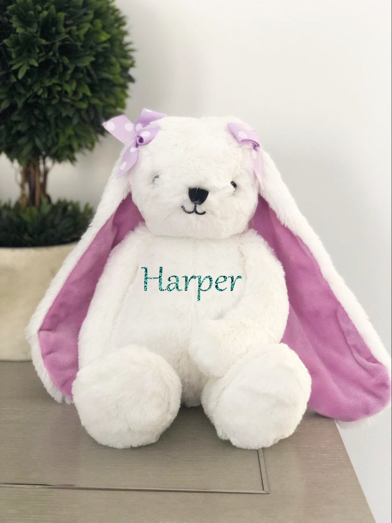 Puede incluir: Un conejito de peluche blanco con orejas largas y un interior morado. El conejito tiene un lazo morado con lunares y el nombre "Harper" bordado en el pecho en color turquesa. El conejito est&aacute; sentado sobre una superficie gris.