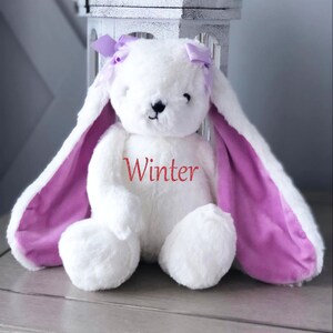 Puede incluir: Un conejito de peluche blanco con orejas largas y un interior morado. El conejito tiene un lazo morado en cada oreja y el nombre "Winter" en escritura roja en el pecho. El conejito est&aacute; sentado frente a una jaula decorativa blanca.