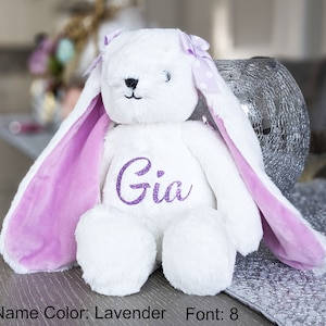 Puede incluir: Un conejo de peluche blanco con orejas largas y un lazo lavanda. El interior de las orejas es lavanda, y el nombre "Gia" est&aacute; bordado con purpurina lavanda en el pecho del conejo. Un peluche lindo y tierno.