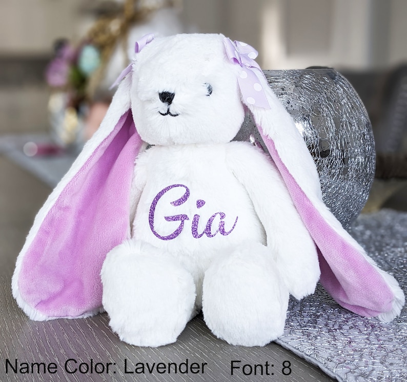 Puede incluir: Un conejo de peluche blanco con orejas largas y un lazo lavanda. El interior de las orejas es lavanda, y el nombre "Gia" est&aacute; bordado con purpurina lavanda en el pecho del conejo. Un peluche lindo y tierno.