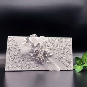 Könnte beinhalten: Eine rechteckige hellgraue Karte mit einem komplizierten geprägten Blumenmuster auf der Oberfläche. Die Mitte ist mit dreidimensionalen Papierrosen, silbernen Blättern und einer durchsichtigen weißen Schleife mit Herzakzenten verziert.