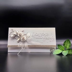 Könnte beinhalten: Eine weiße, rechteckige Hochzeitsgrußkarte mit dem geprägten Text On Your Wedding Day. Die Karte ist mit zwei weißen Papierrosen, silbernen Blättern, einer transparenten weißen Schleife, einem glitzernden horizontalen Band und einem kleinen Herz.