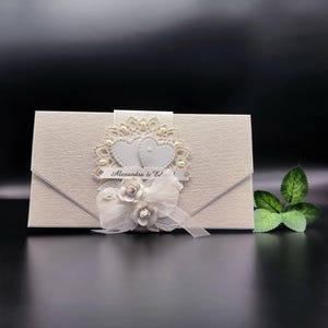 May include: Ivory wedding invitation with a floral embossed pattern. Features a decorative embellishment of two white hearts, lace, and a ribbon with the text "Alexandra & Ed". A white bow and flowers add to the design.