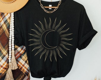 May include: Black t-shirt with a gold sun and moon graphic design. The sun has rays and the moon is a crescent shape.