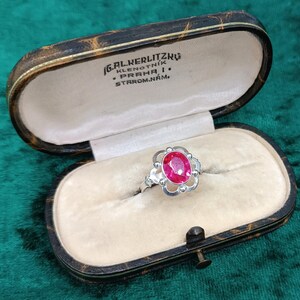 Puede incluir: Un anillo de plata con una gema rosa brillante, ovalada y grande, engastada en un diseño floral decorativo. El anillo se muestra dentro de un joyero vintage de cuero con texto en la tapa.